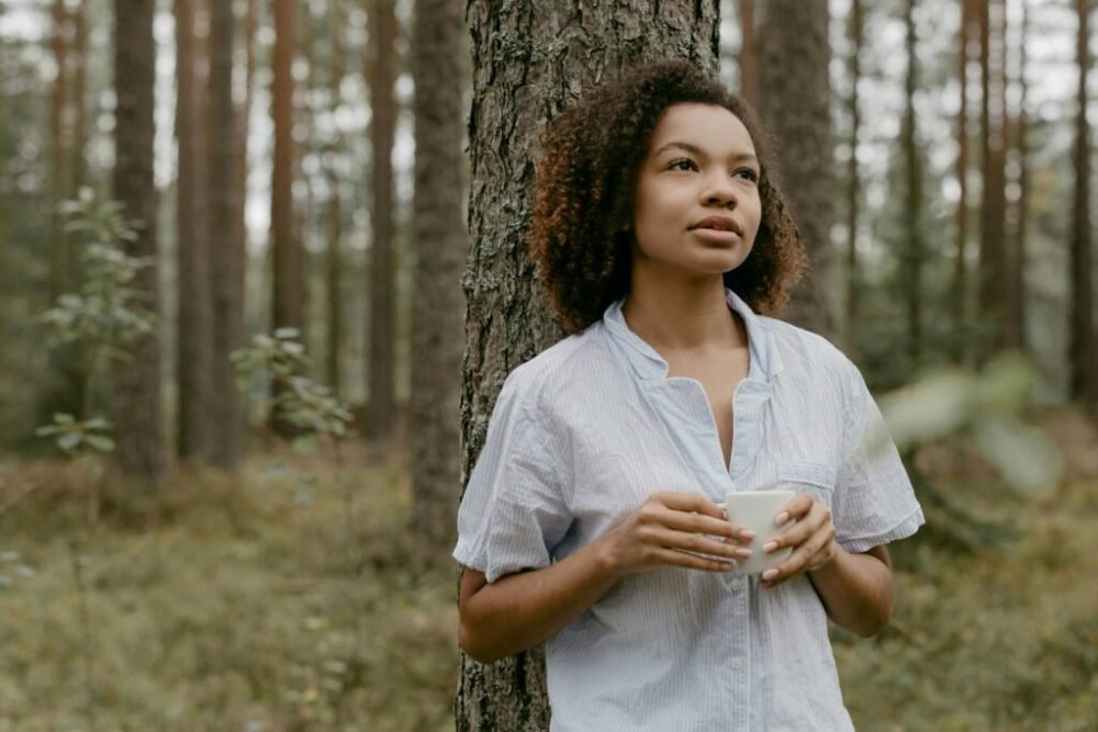 A woman enjoys a peaceful moment with a mug in a serene forest setting, embodying relaxation.